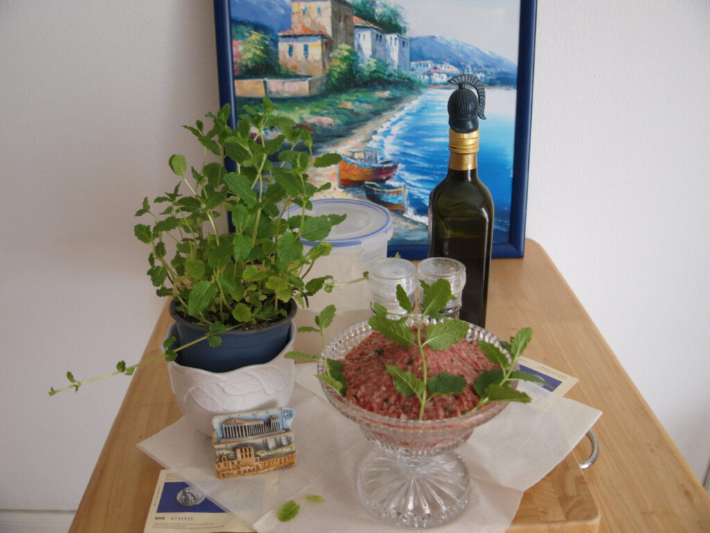 Minze Fleischbällchen griechisch in Glasschale präsentiert mit griechischer Landschaft als Bild im Hintergrund und Minzetopf zur Untermalung